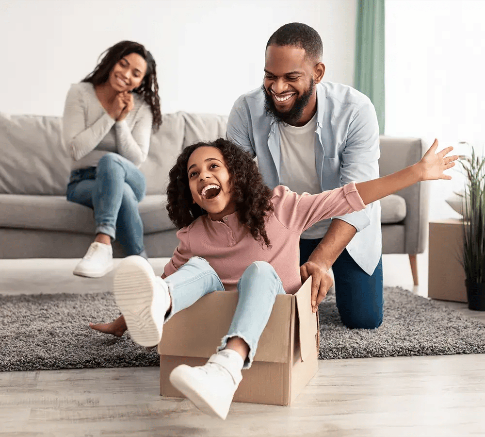 A mom on the couch smiles as her husband pushes their daughter around on the floor, and she laughs as she sits in a moving box with her arms outstretched.