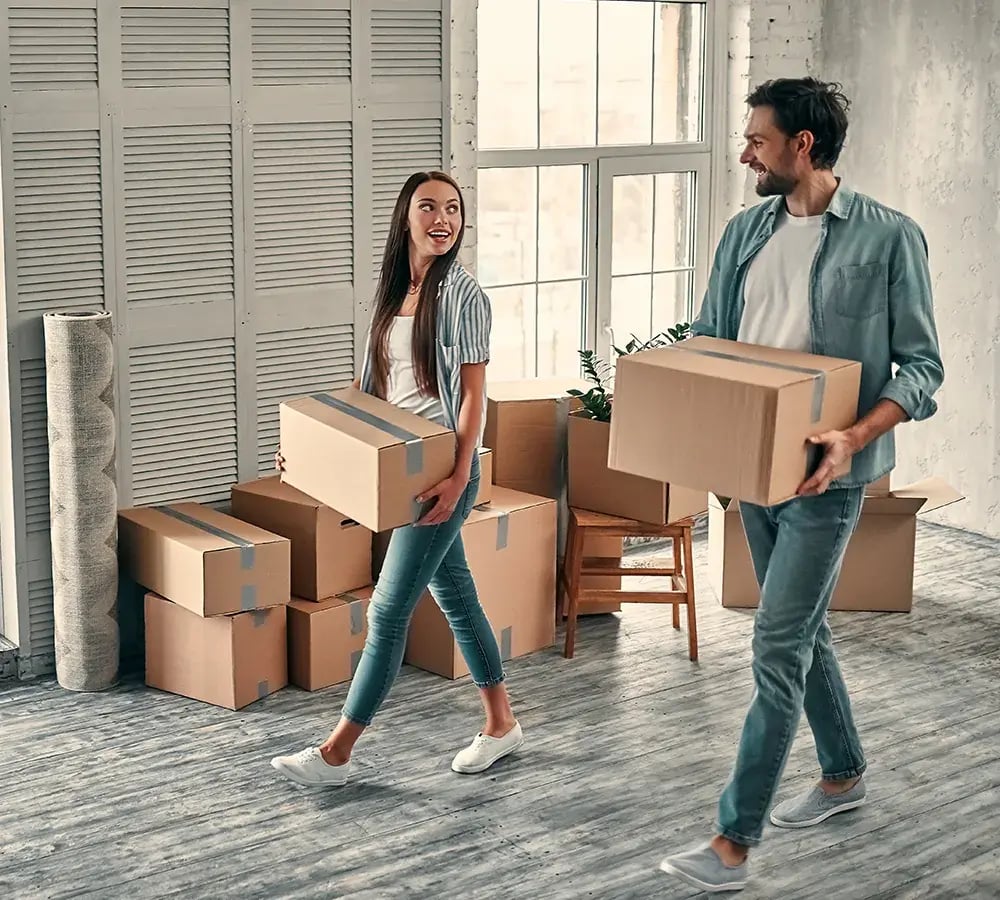 A couple are happily carrying moving boxes through a room in a house they appear to be moving into
