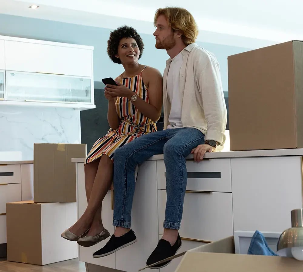 A couple, sitting on the counter looking satisfied and surrounded by moving boxes in the room
