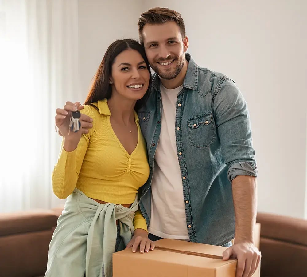 A couple standing by a moving box, with smiles on their face and house keys in hand