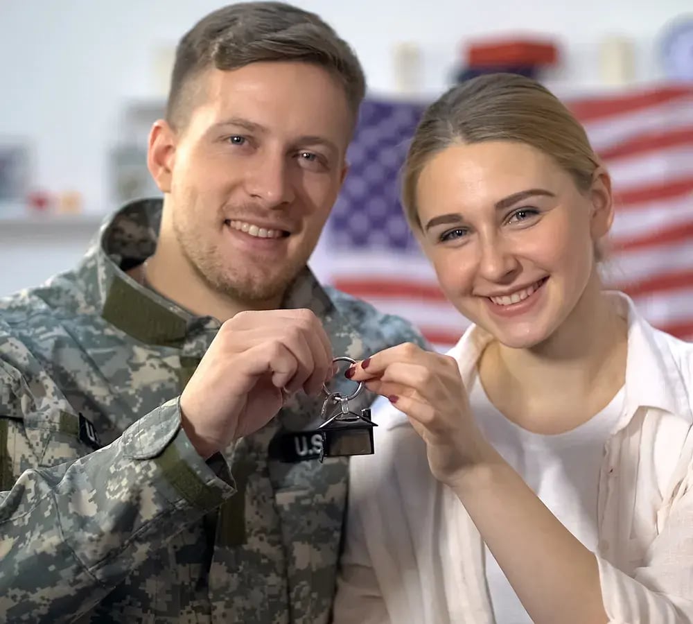 A couple smiling at the camera, each with one hand holding up a key, with an American flag in the background.