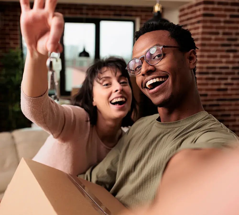 Happy couple in a brick house, one holding a moving box and hone holding up house keys