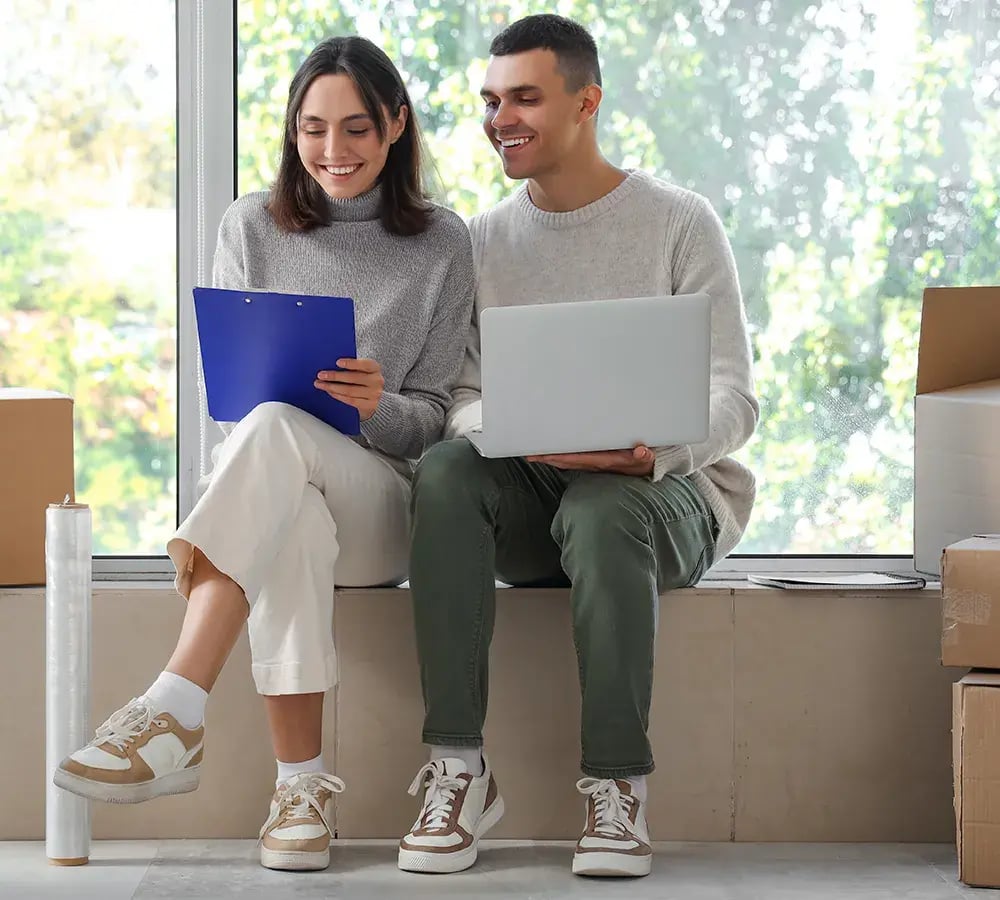 A happy couple sits amongst their moving boxes, smiling and looking over notes on a laptop and a clipboard