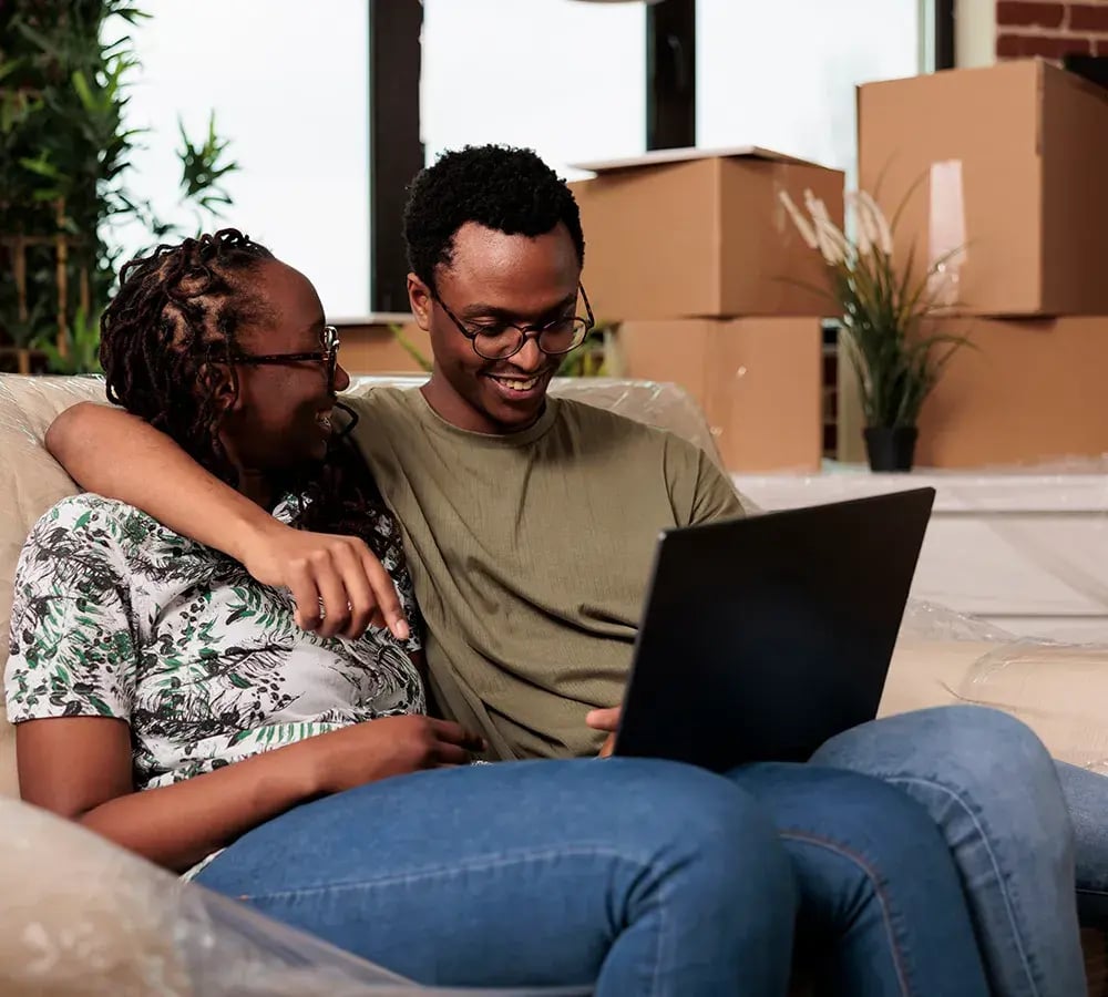 A happy couple sitting on a couch and laughing together, with a laptop in front of them and moving boxes in the background