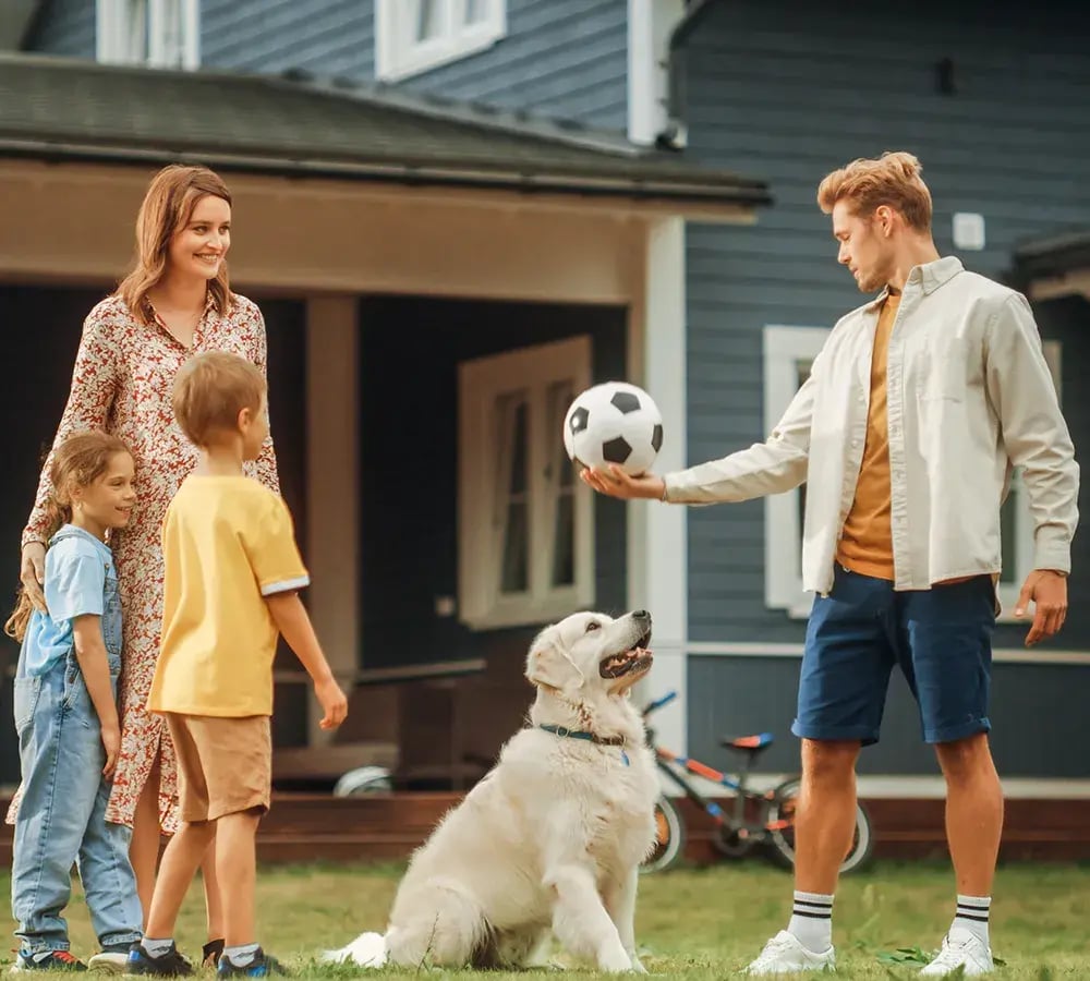 A family of four, with their dog, laughing and playing with a soccer ball